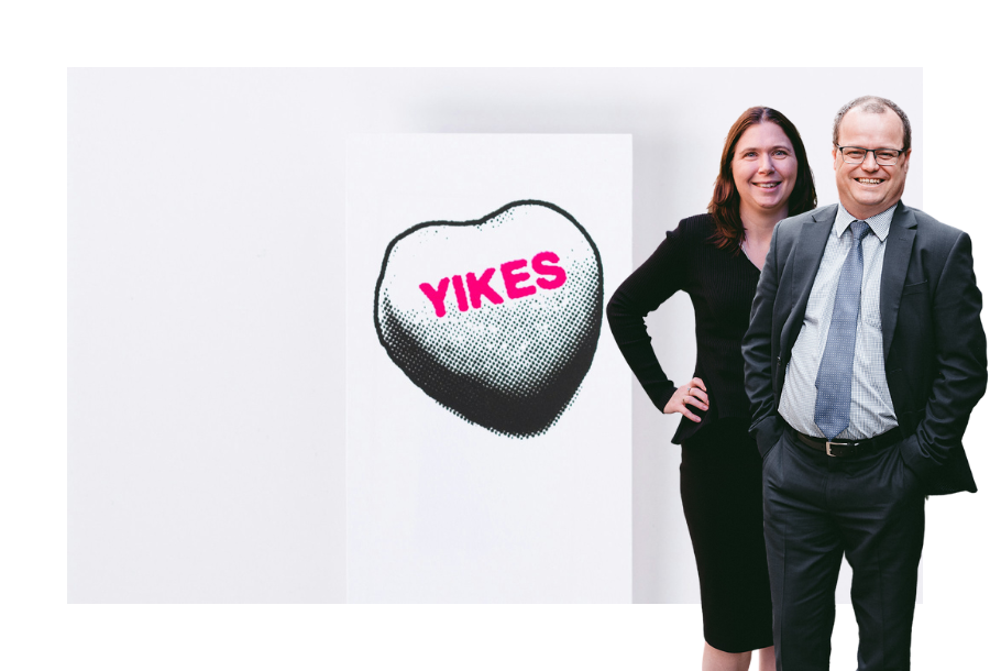Pictured, Adam Wightman and Karlene Wightman standing smiling, behind then is an image of a white heart shape with the word "yikes" written in pink.