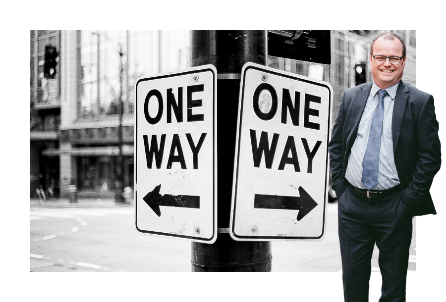 Pictured: Adam Wightman standing smiling wearing a black suit with a light blue shirt and blue tie with his hands in his trouser pockets. Behind him is an image of two "One Way" road signs next to each other pointing in opposite directions.