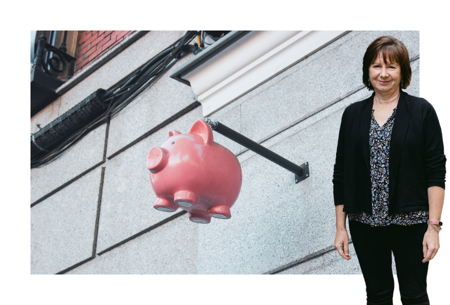 Pictured: Nancy Robertson standing smiling in a black cardigan with a floral shirt underneath and black trousers. Behind her is an image of a pink piggy bank handing from a pole coming outward from an exterior building wall.